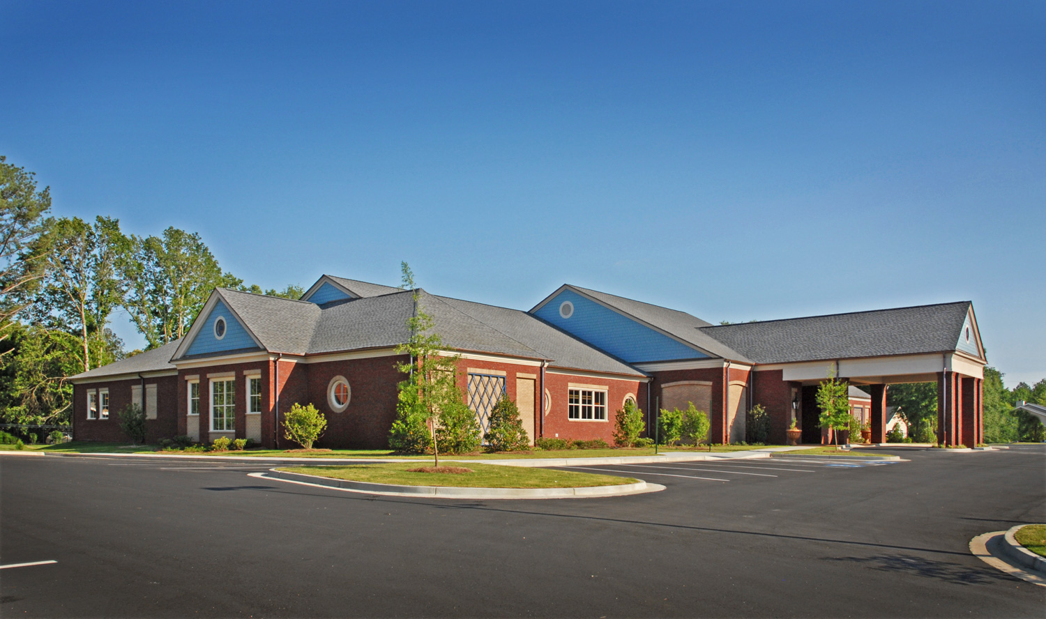 A single-story brick building with a gray roof sits amid greenery by an empty parking lot under a clear blue sky.