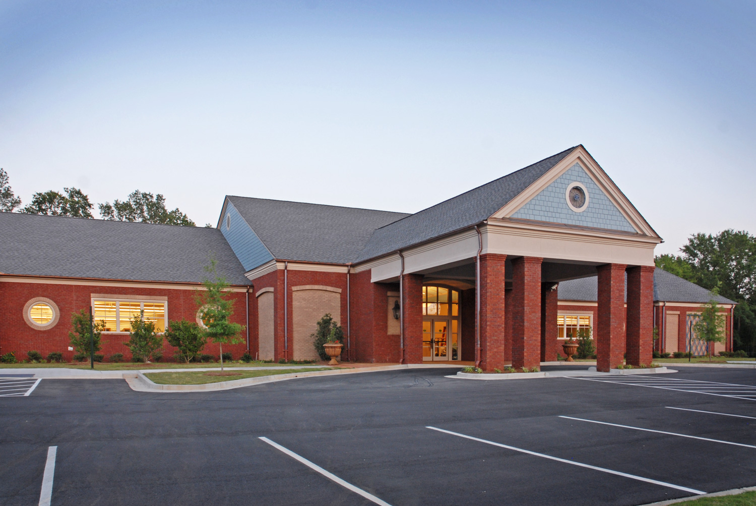 A modern red brick building with front columns and tall windows stands under a clear sky, surrounded by landscaping and an empty lot.