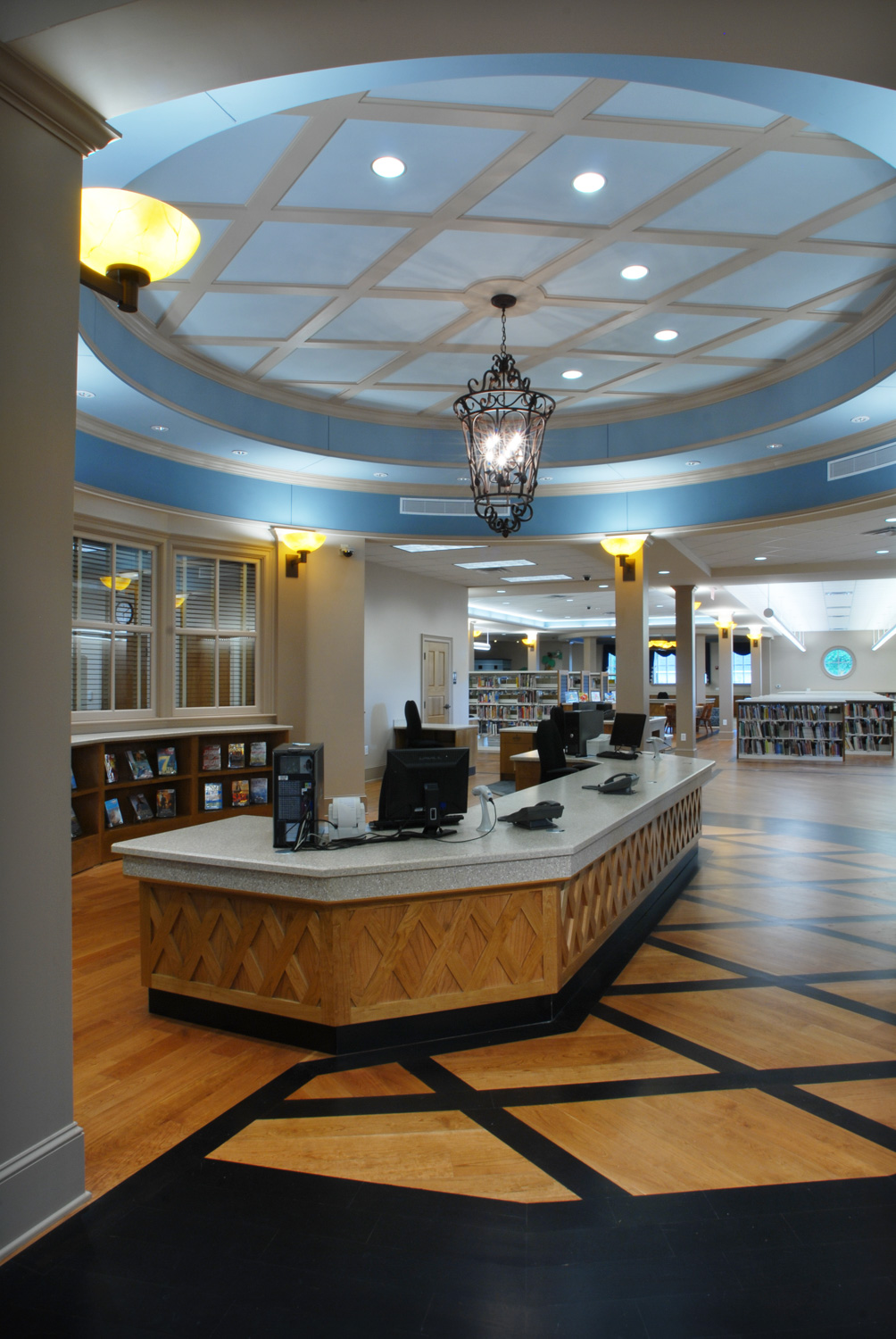 A spacious modern library interior with a curved wooden check-out desk, decorative ceiling light, bookshelves, and patterned floor.