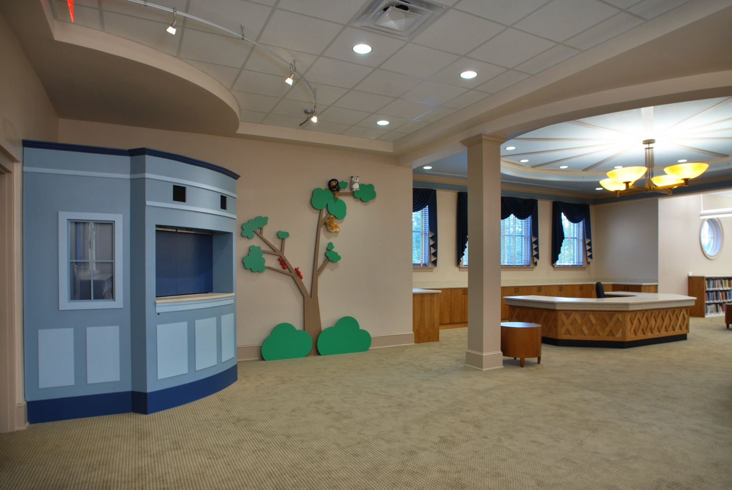 Spacious, well-lit room with a light blue puppet theater, tree wall decor, carpeted floor, large desk, and dark blue curtained windows.