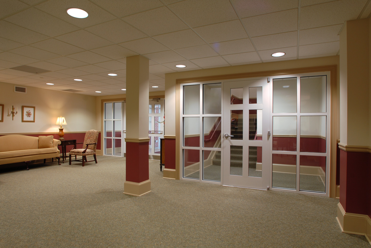 A carpeted hallway with beige walls, red trim, ceiling lights, a sofa set on the left, and glass doors to stairs on the right.