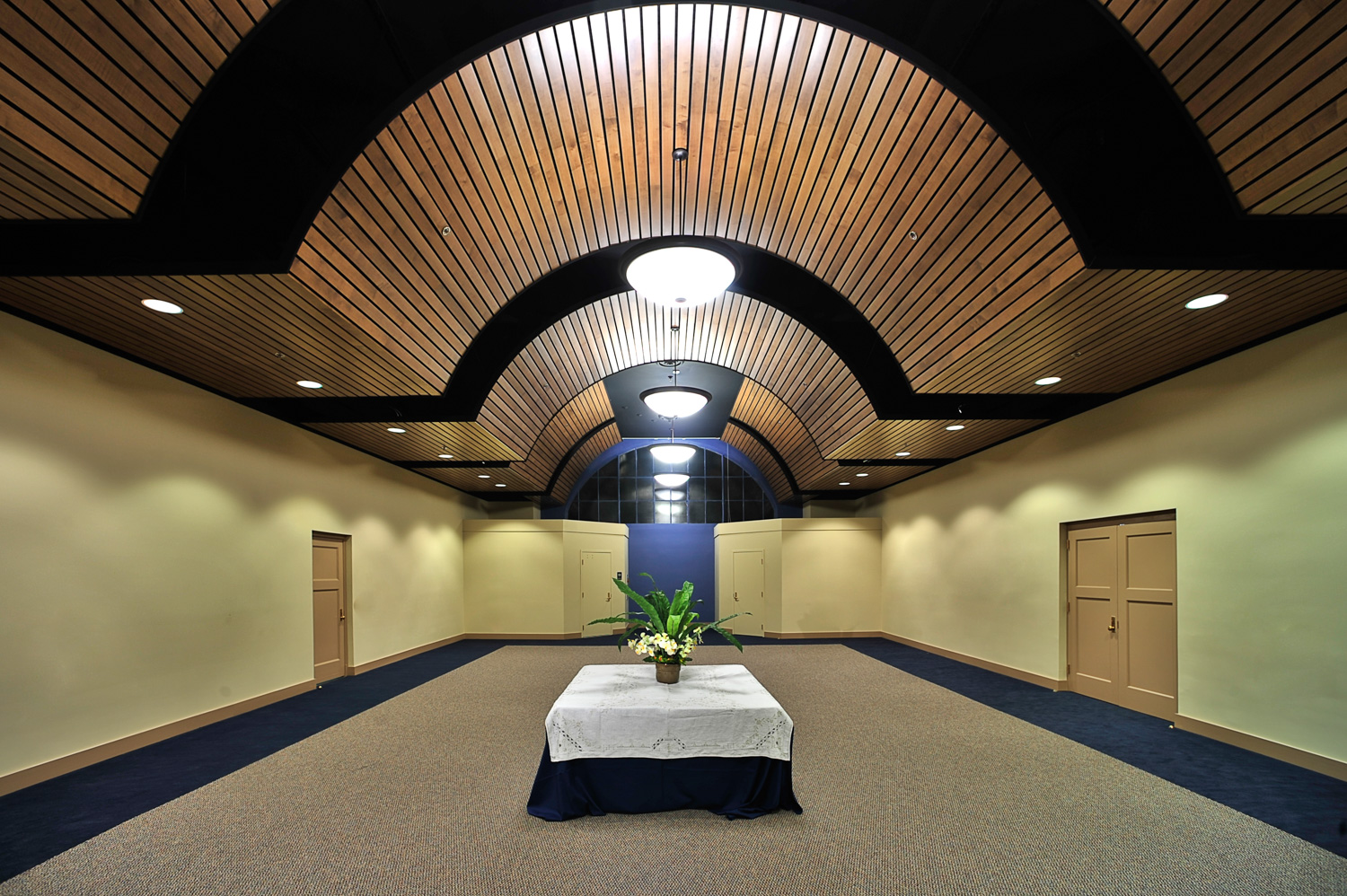 Symmetrical, well-lit hallway with curved wooden ceiling, beige walls, blue carpet borders, table with flowers, and doors on both sides.