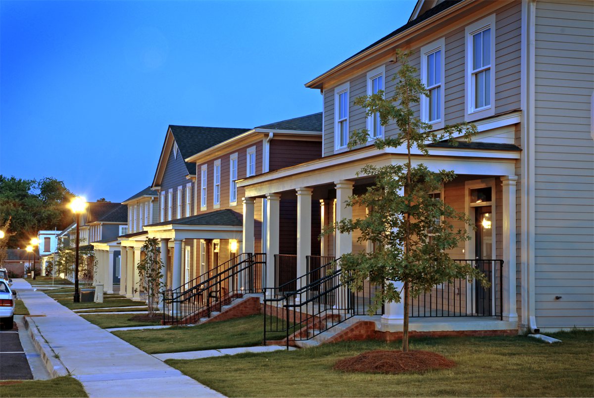 Modern two-story suburban houses with front porches and neat lawns at dusk, streetlights illuminating a clean sidewalk.