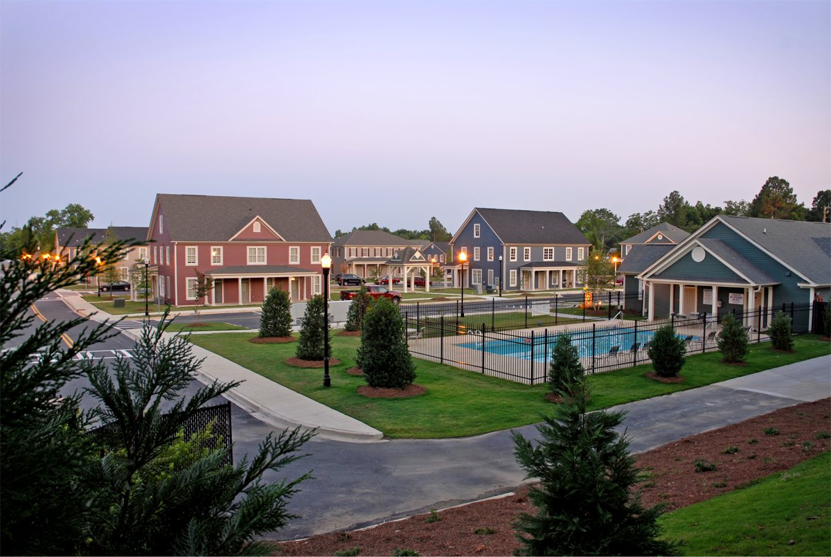 Suburban neighborhood at dusk with colorful two-story houses, landscaped lawns, a pool, sidewalks, and small street trees.