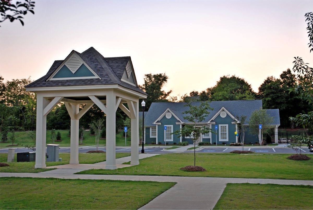 A small blue building with white trim stands behind a pavilion in a grassy park, sidewalk, trees, empty parking spots, sunset sky.