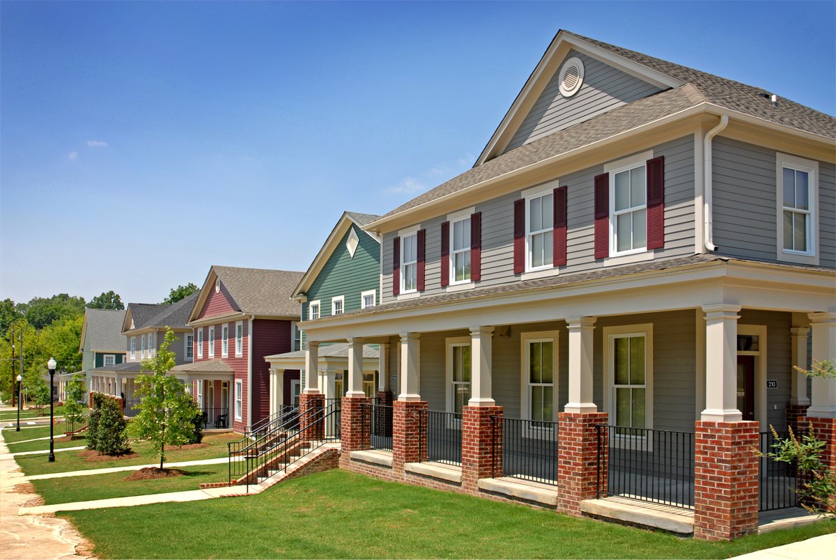 Modern suburban houses with colorful exteriors and front porches line a tidy grassy street beneath a clear blue sky.
