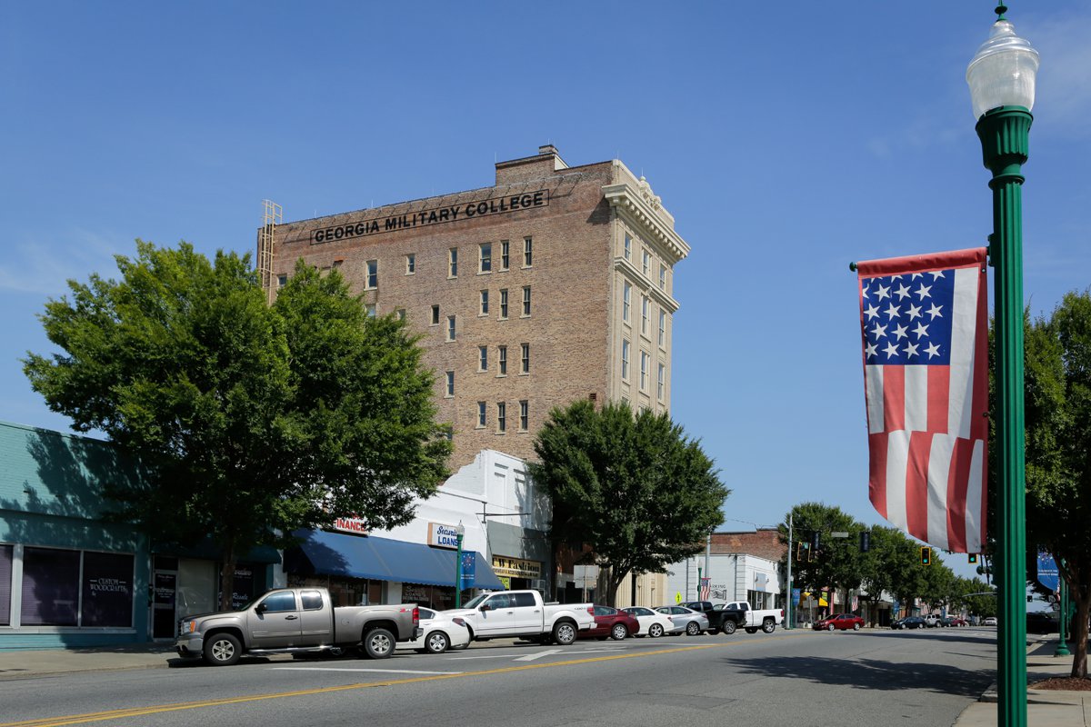 A small town street with parked cars, storefronts, trees, a tall building labeled Georgia Military College, and an American flag banner on a lamppost. The sky is clear and blue.