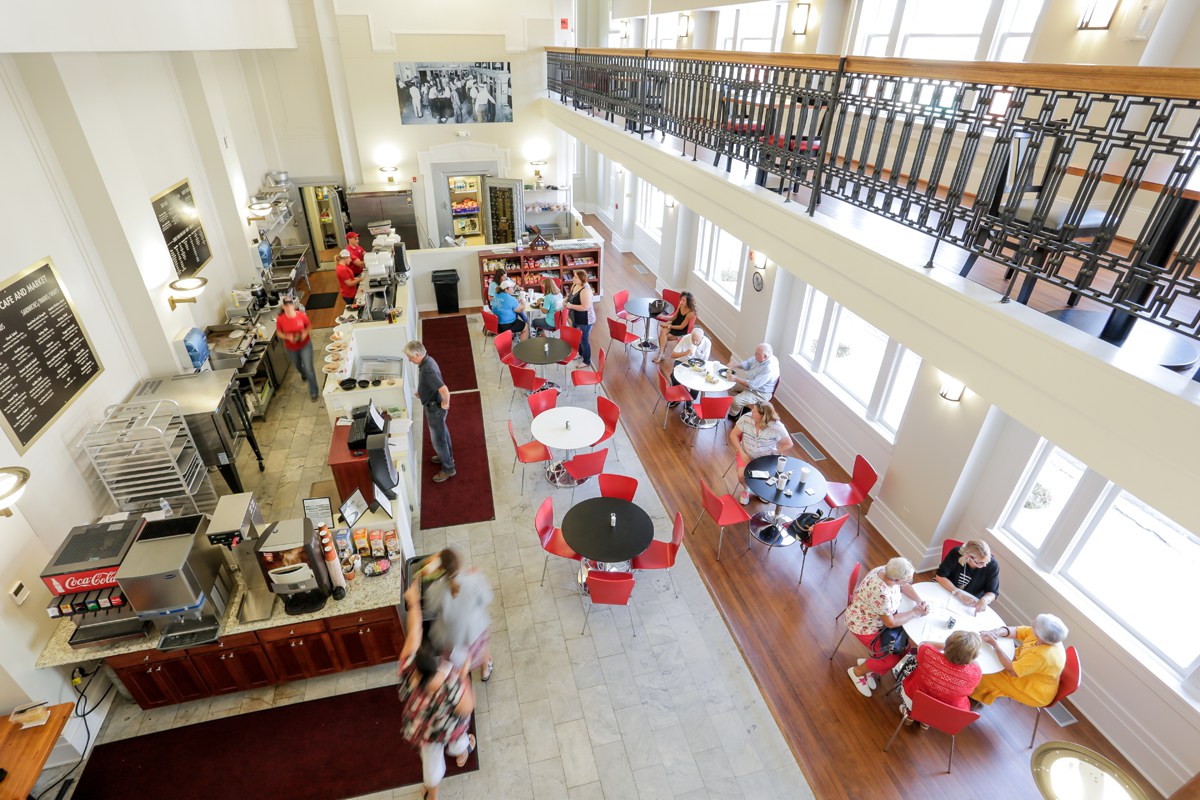 Bright, modern café with red chairs and white tables viewed from above; people seated, staff behind counter, natural light streams in.
