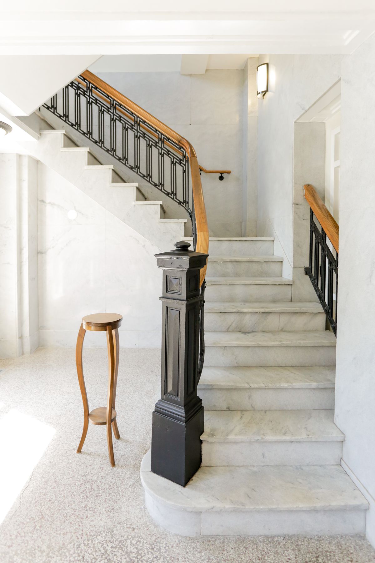 A marble staircase with black and wood handrail, white walls, and a small wooden side table in a bright sunlit interior.