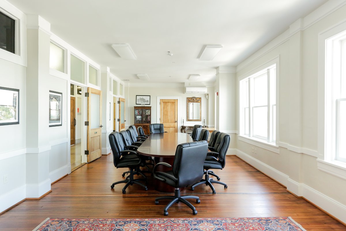 Bright conference room featuring a long oval table, black office chairs, wooden floors, large sunlit windows, and framed wall art.
