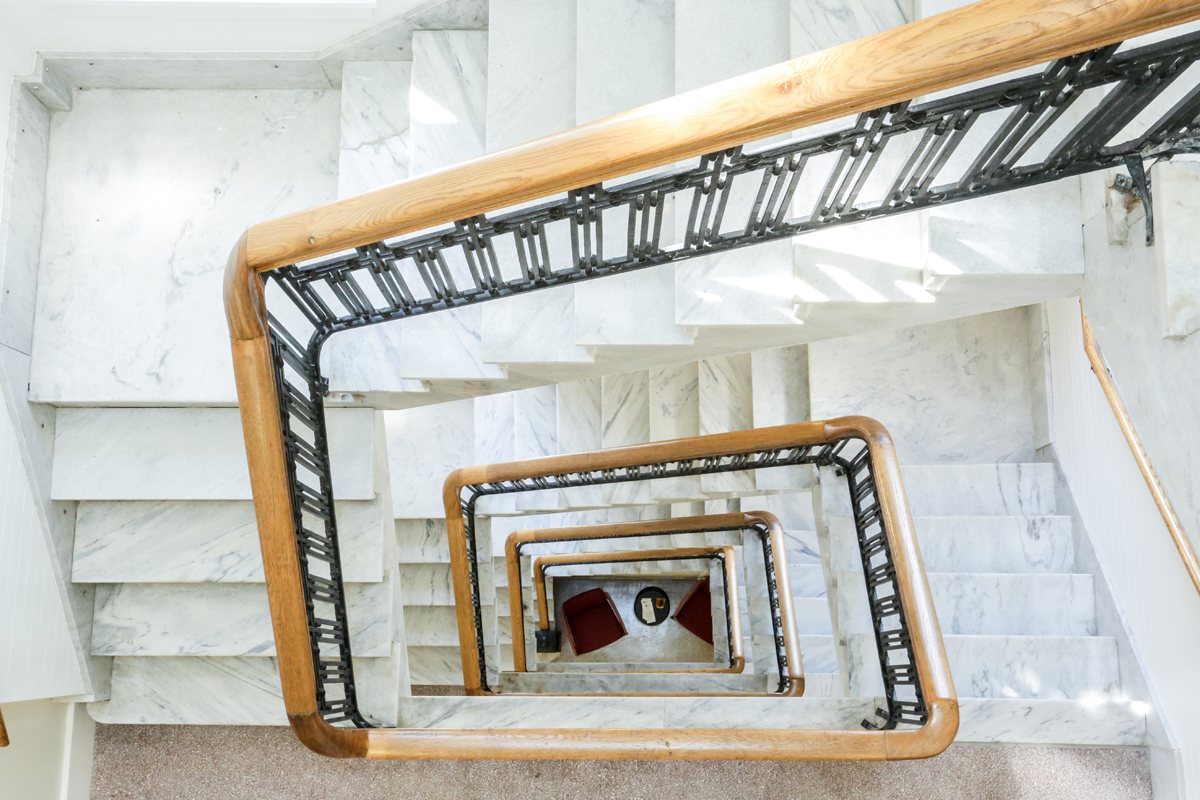 Aerial view of a spiral white marble staircase with wood and black metal railing; two red chairs and round table below on marble floor.