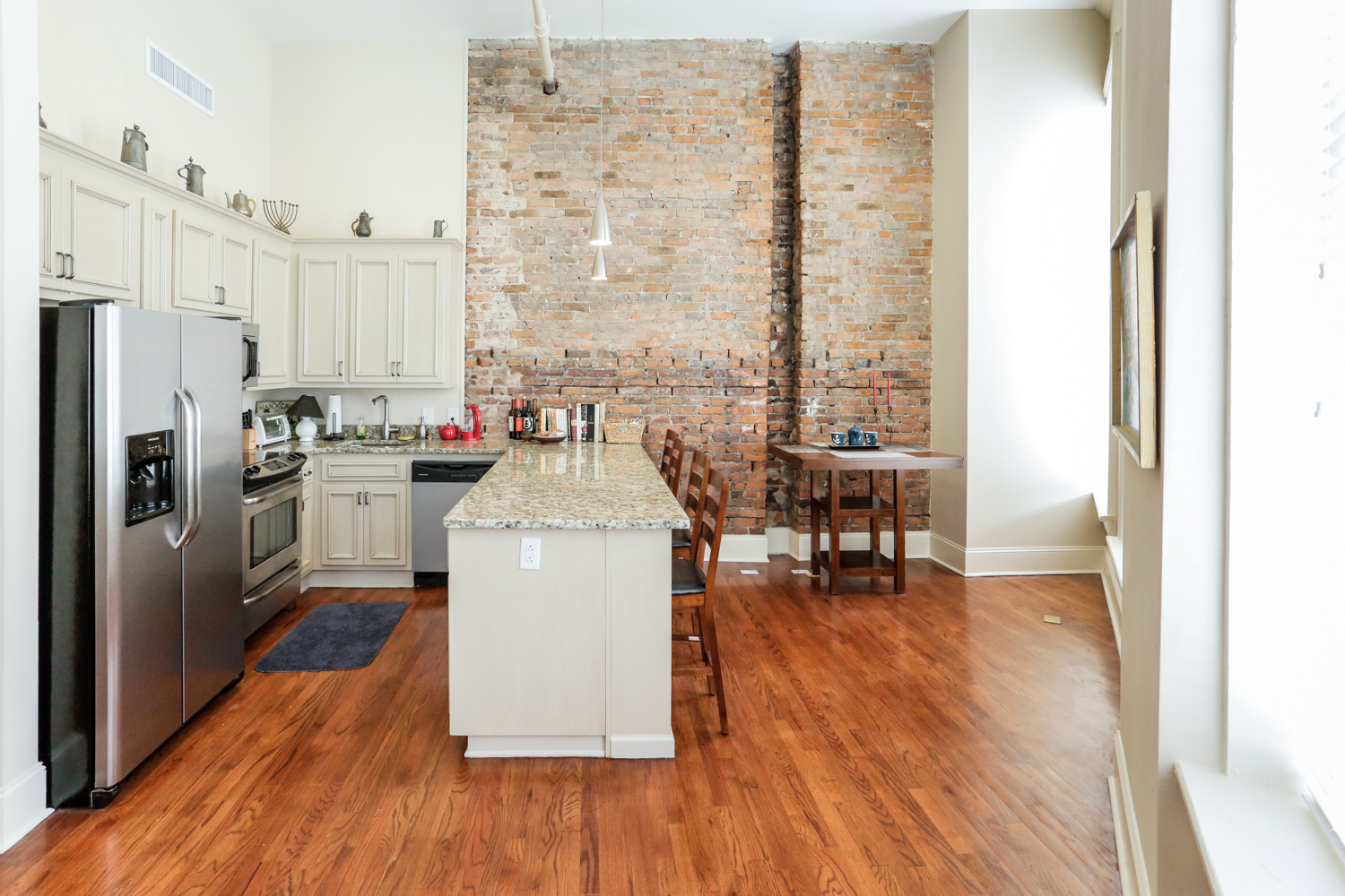 A modern kitchen with light cabinets, stainless steel fridge, granite island, wooden chairs, exposed brick, and hardwood floors.