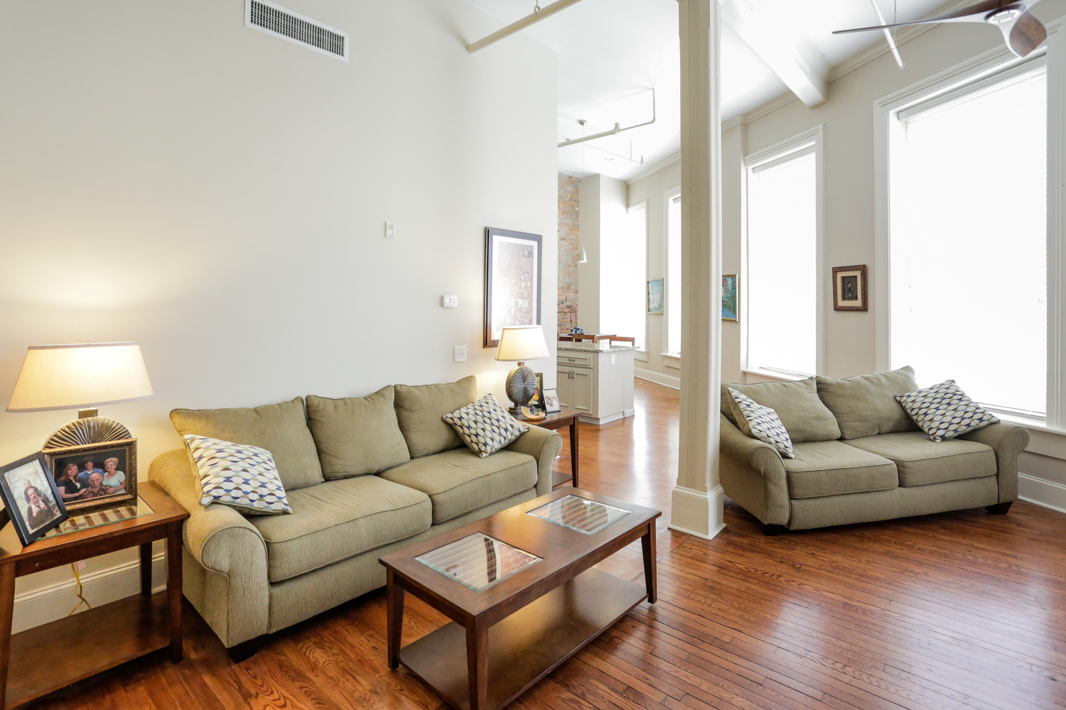 Bright living room with hardwood floors, beige sofas, patterned pillows, glass coffee table, side table with lamps, large windows.