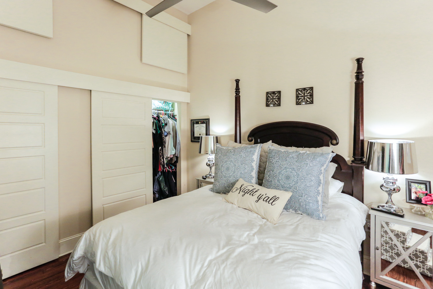 A cozy bedroom with a four-poster bed, white bedding, blue decorative pillows, and a Night y'all pillow. Two mirrored nightstands with lamps sit beside the bed, and an open closet is visible in the corner.