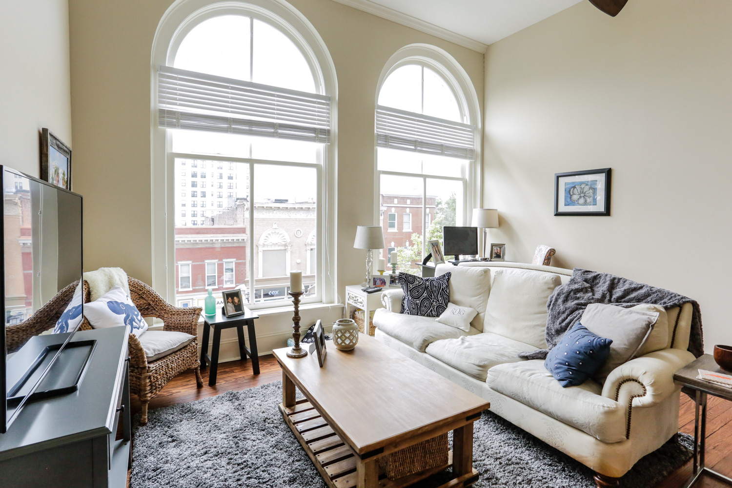 Bright living room with large arched windows, white sofa, wicker chair, wood coffee table, decor accents. City buildings outside.