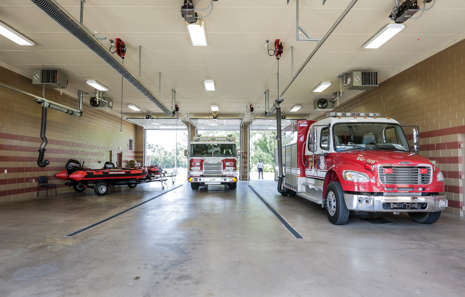 A fire station garage with two fire trucks and a red inflatable rescue boat on a trailer, parked in open bays with daylight coming in.