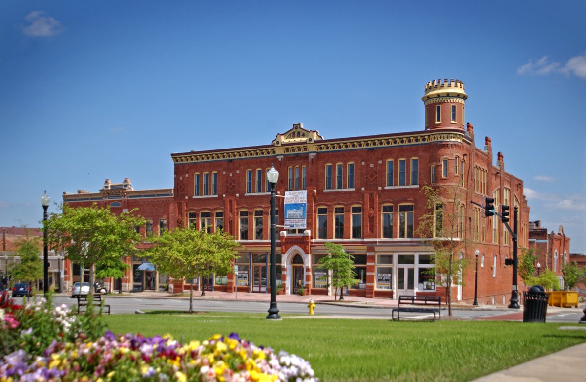 Historic red brick building with a small turret on a sunny street corner, trees, benches, and colorful flowers under blue sky.