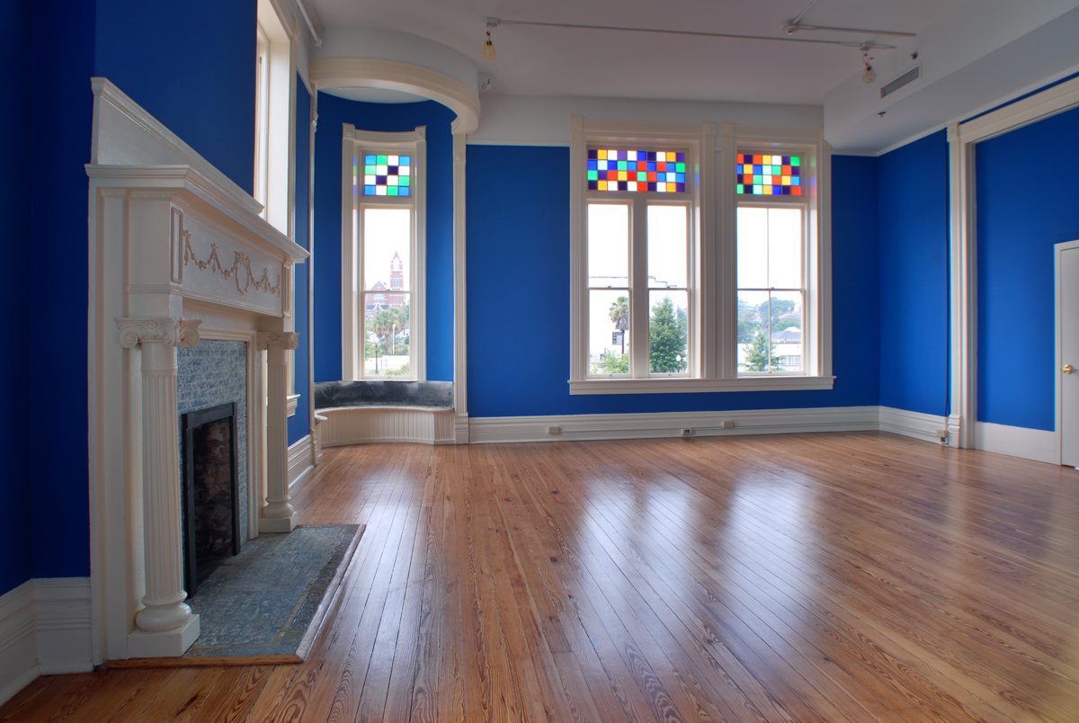 Spacious empty room with bright blue walls, large windows topped with colorful stained glass, white-mantled fireplace, wood floors.