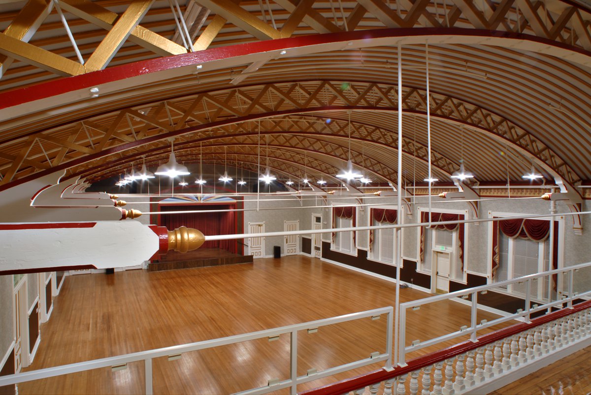 Spacious empty ballroom with polished wood floors, high arched beamed ceilings, red curtained windows, bright lights, and a stage.