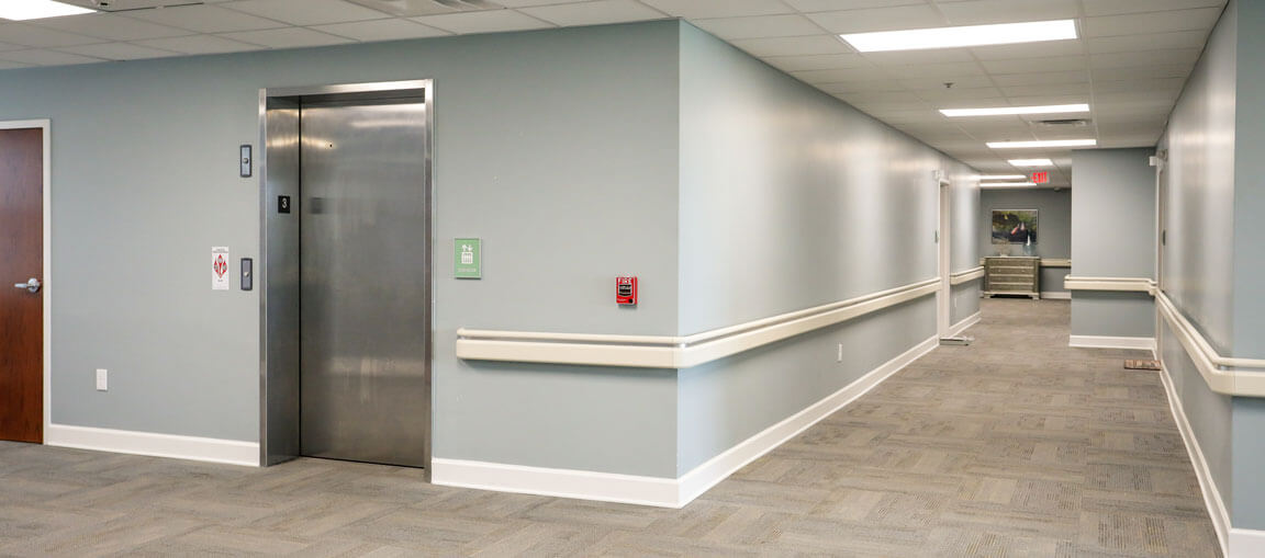 A clean empty hallway with light gray walls, carpeted floors, closed elevator doors on the left, handrails, and ceiling lights.