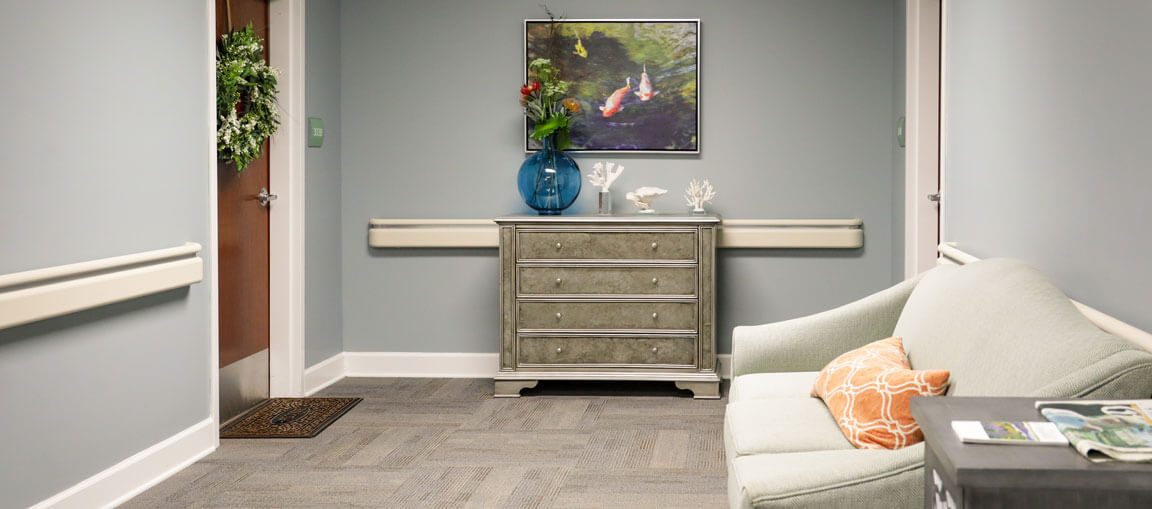 Cozy waiting area with a light green sofa, orange pillow, magazines on a small table, gray dresser, fish painting, and door with wreath.