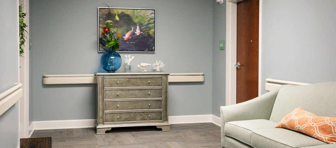 A hallway with light blue walls, silver dresser with decor, painting above, green couch with orange pillow, closed wooden door behind.
