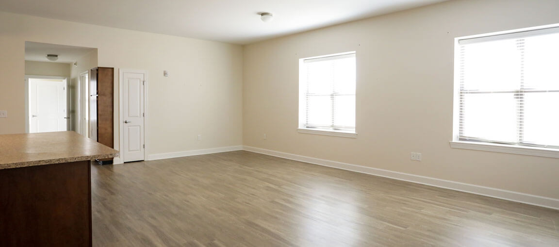 A spacious empty room with light wood floors, beige walls, large windows with blinds, and an open kitchen area. Natural light fills the space.