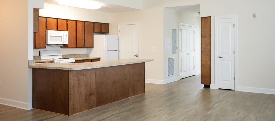 Modern kitchen with wooden cabinets, white appliances, large island, light wood floors, and hallway with white doors and neutral walls.
