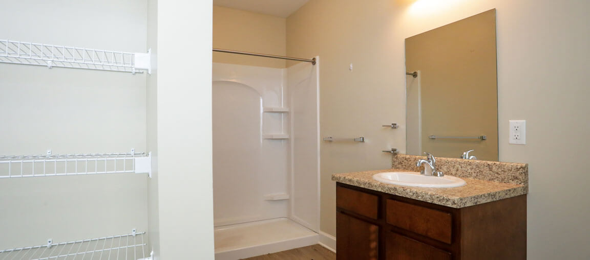 A bathroom with a walk-in shower, single sink vanity with granite countertop, large mirror, wire shelves on left wall, light beige walls.