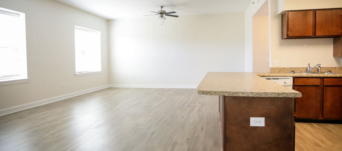 Empty apartment with light wood floors, beige walls, large windows, ceiling fan; kitchen has brown cabinets and a dishwasher.