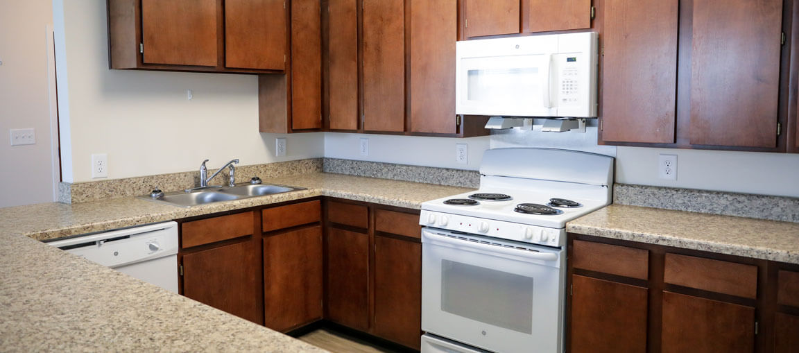 A kitchen with brown cabinets, beige granite counters, a white stove and microwave, double sink, dishwasher, and light walls.