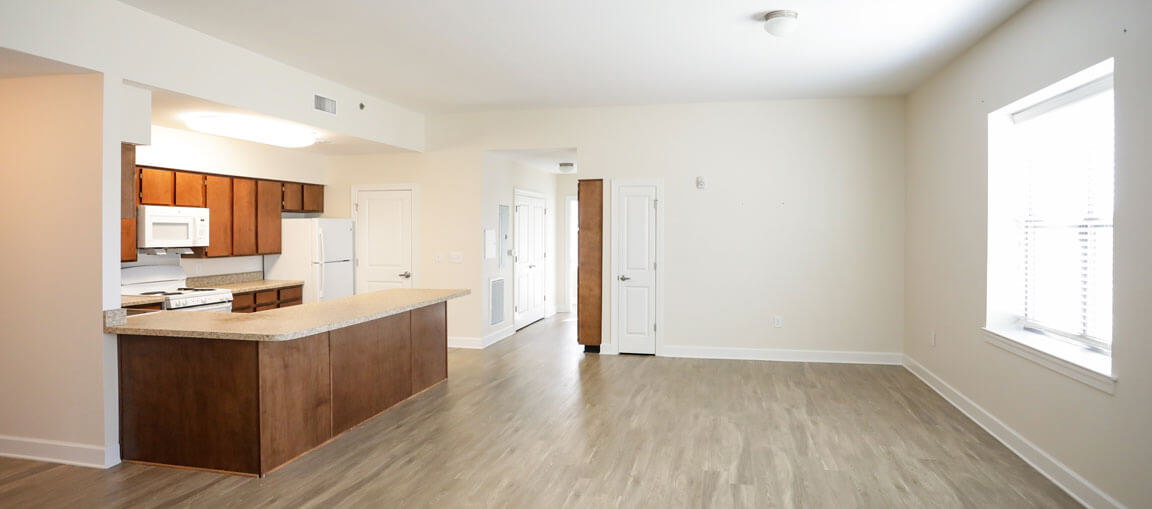 Spacious, empty apartment with light wood floors, white walls, brown kitchen cabinets, white appliances, large counter, and window.