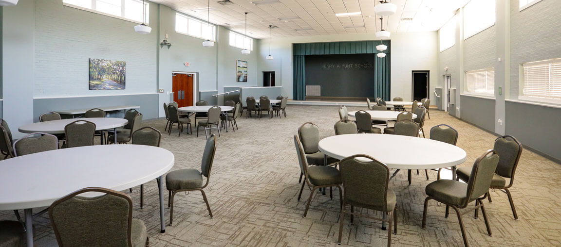 Spacious multi-purpose room with round tables and chairs, carpet, stage at the end, large windows, artwork, and bright ceiling lights.