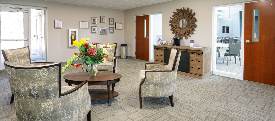 Bright, modern waiting area with patterned armchairs, coffee table and flowers, wood sideboard, mirror, photos, and doors.