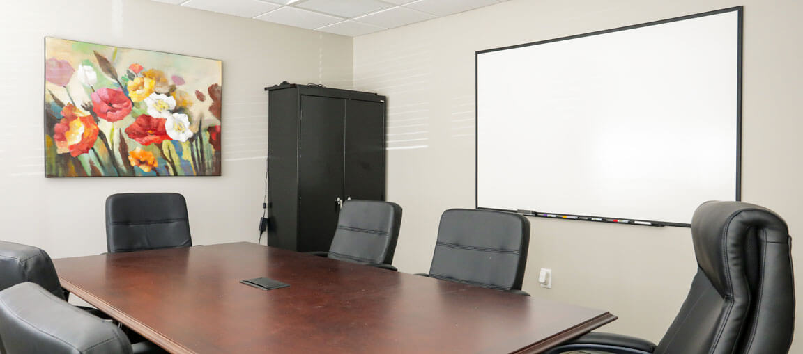 Conference room featuring a wooden table, five black chairs, whiteboard, black storage cabinet, and a vibrant floral painting on the wall.