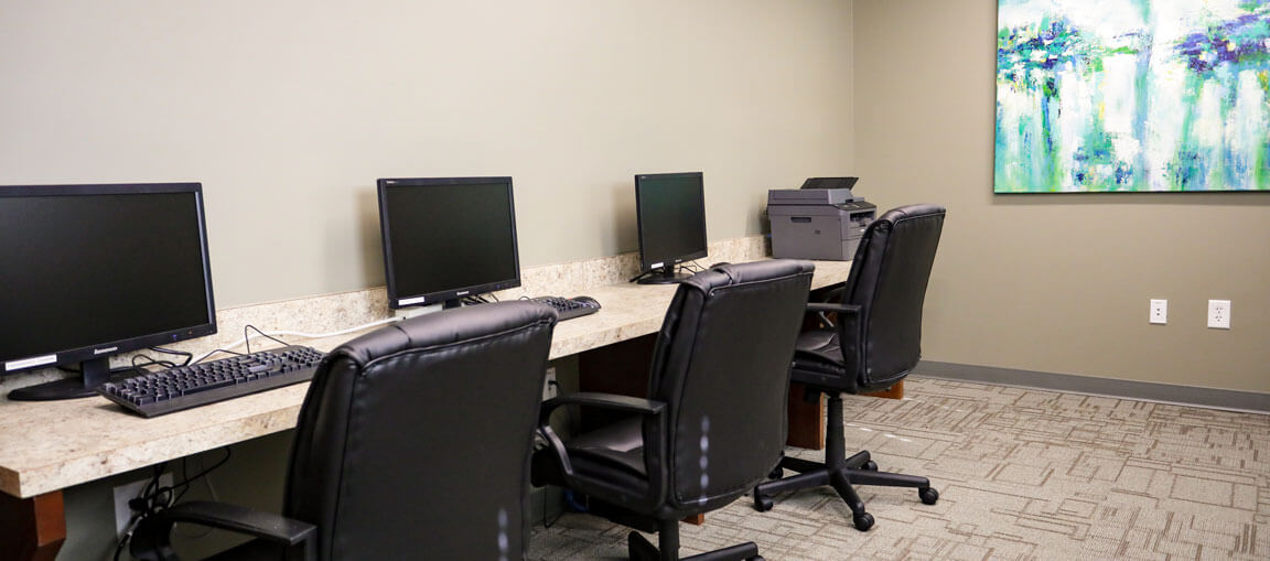 Three computer workstations with black chairs on a long counter, plus a printer and abstract art in a modern carpeted office.