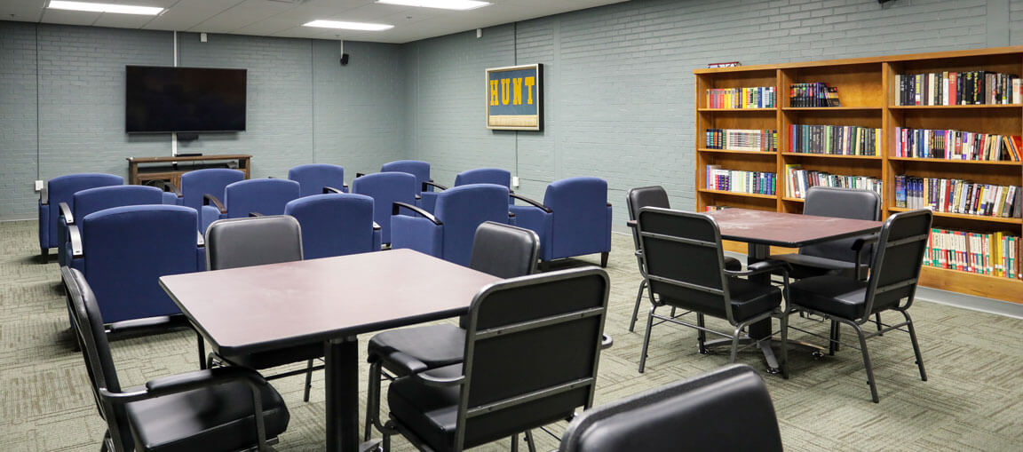A classroom with blue chairs facing a TV, tables and black chairs on one side, bookshelf of books, and framed art on a gray wall.