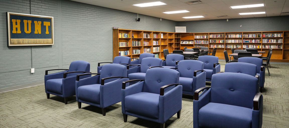 A library room with rows of blue armchairs facing forward, bookshelves filled with books, a framed HUNT sign, and tables in back.