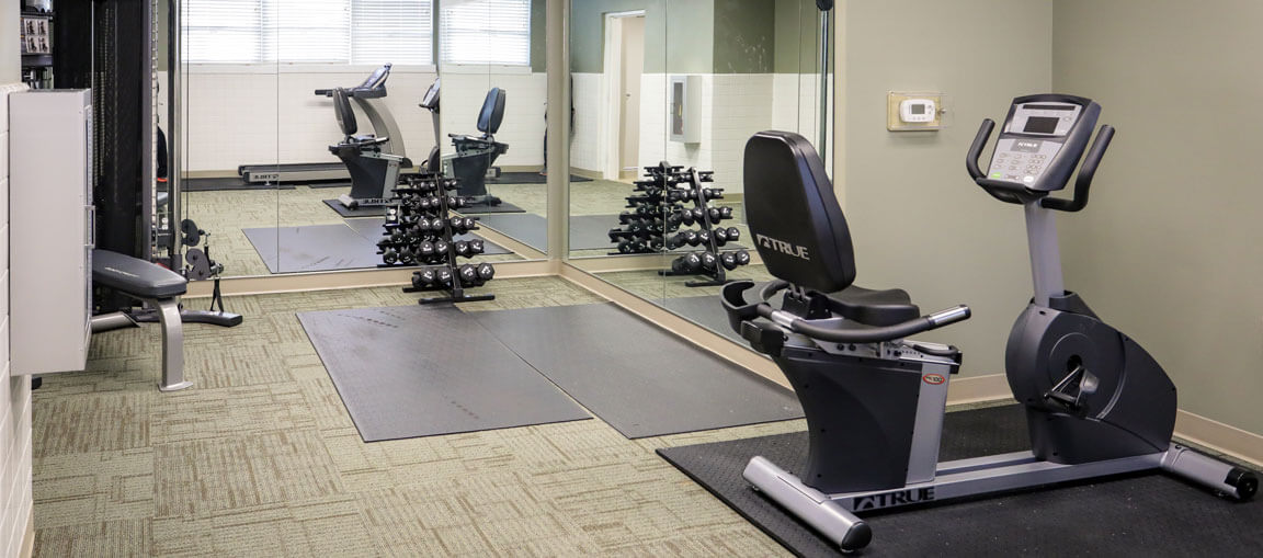 A small gym room with a recumbent bike, weight bench, dumbbells on a rack, and wall mirrors. Light green walls and carpeted floor.