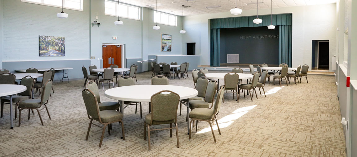 A spacious multipurpose room with round tables, green chairs, beige carpet, green walls, hanging lights, wall art, and a small stage.