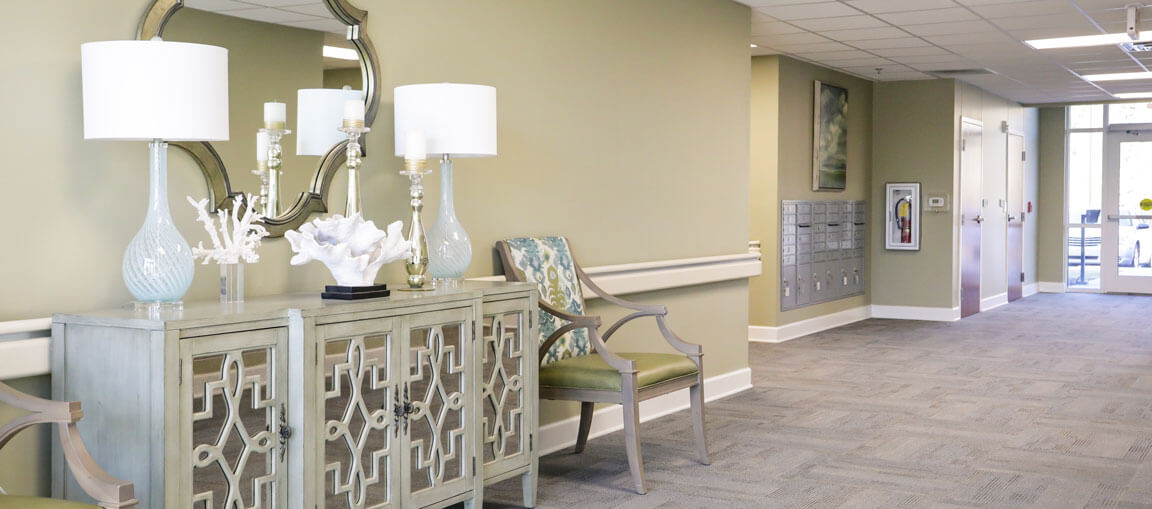 Hallway with pale green walls, gray carpet, console table with lamps, decorative items, patterned chair, and a round mirror.