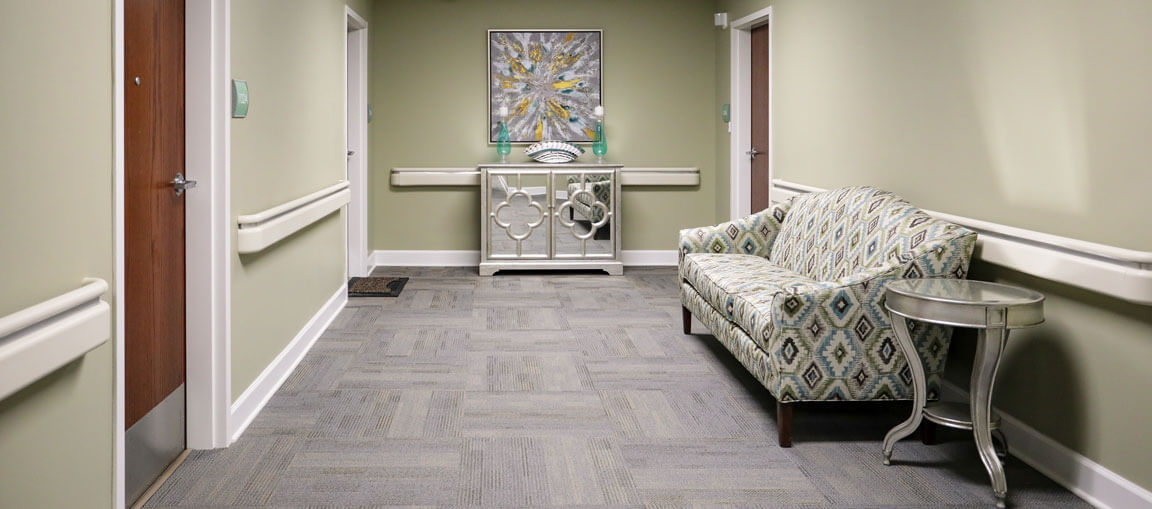 Hallway with light green walls, gray carpet, patterned sofa, small round table, white console table and painting at the end; wooden doors on each side.