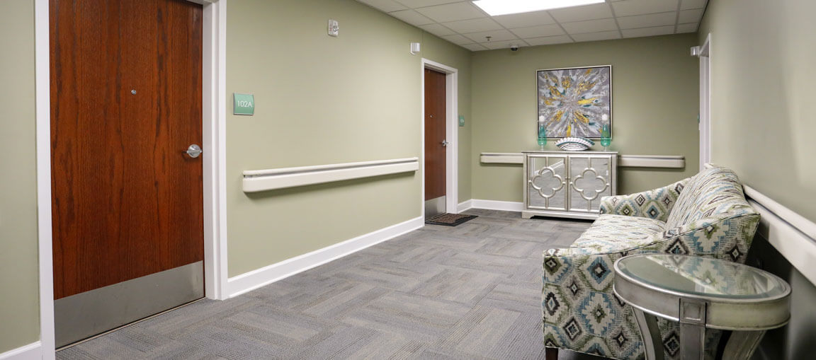 A hallway with light green walls, grey carpet, a wooden door on the left, patterned couch, glass side table, console table, and abstract painting.