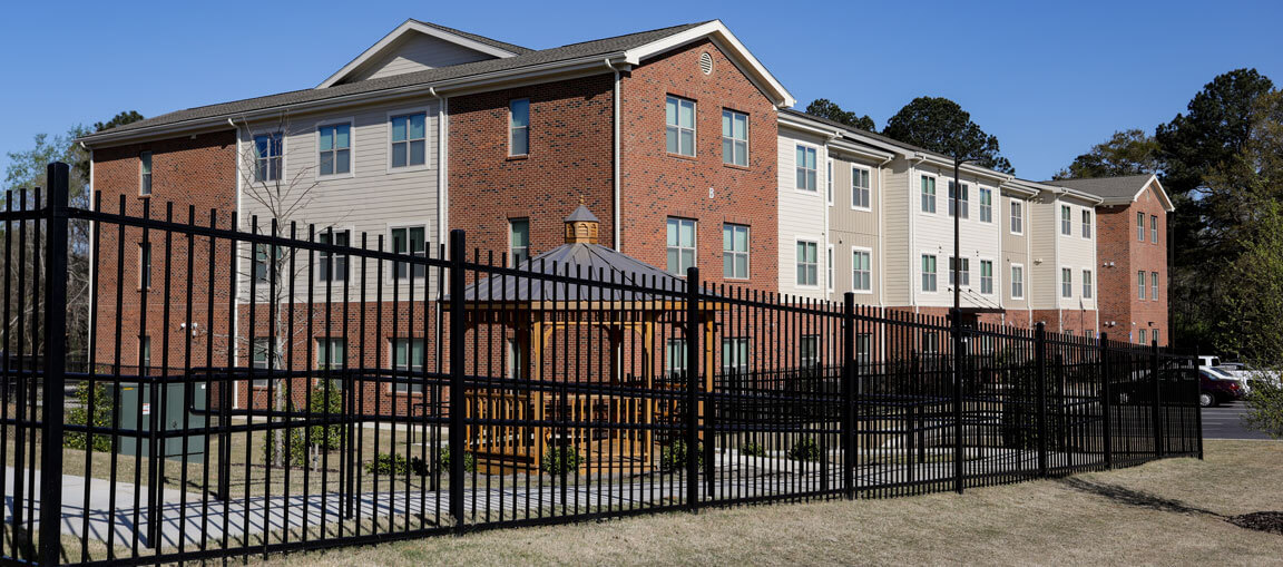 Three-story apartment building with red brick and beige siding behind a black fence, gazebo in yard, trees and blue sky behind.