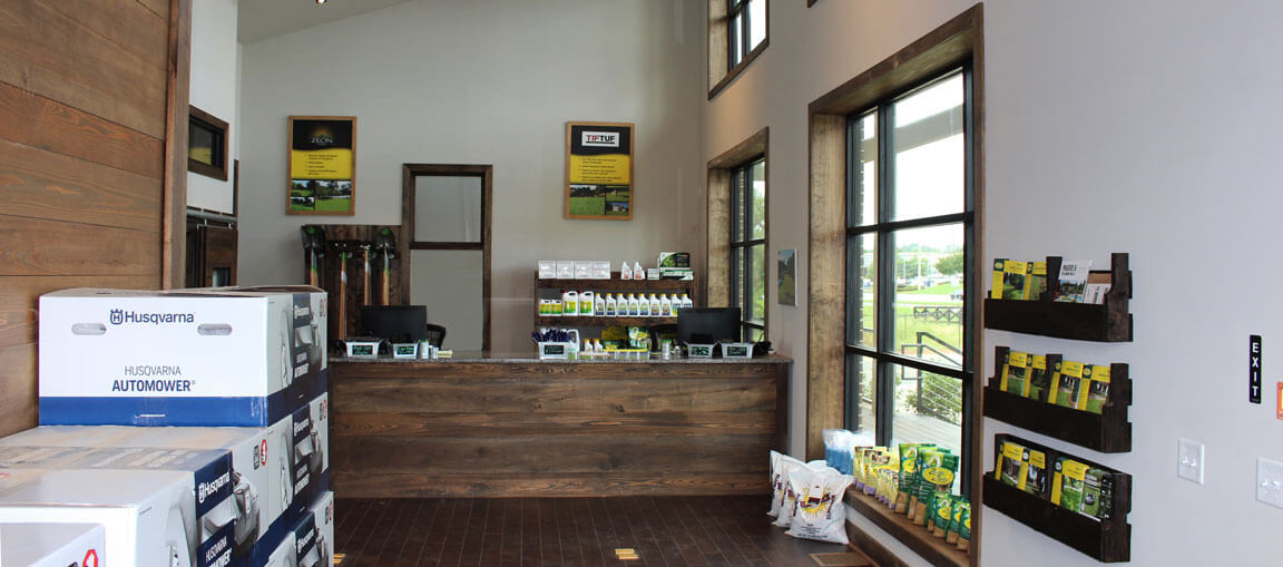 A garden supply store with a wooden counter, product displays, mulch and seed bags, tools on the wall, and large sunlit windows.