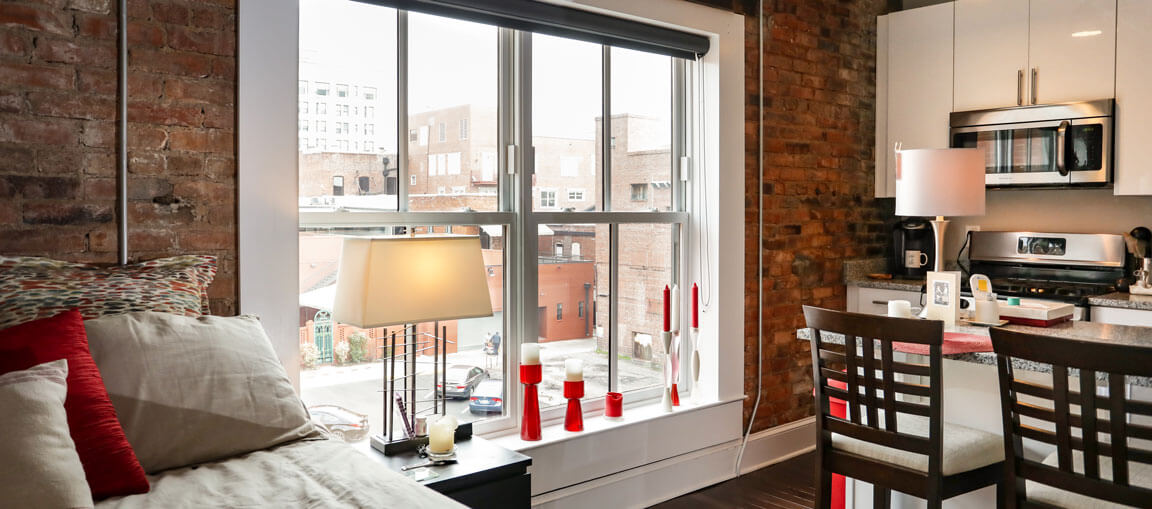 Cozy apartment with exposed brick, red and patterned pillows on the bed, large city-view window, modern kitchen with red accents.