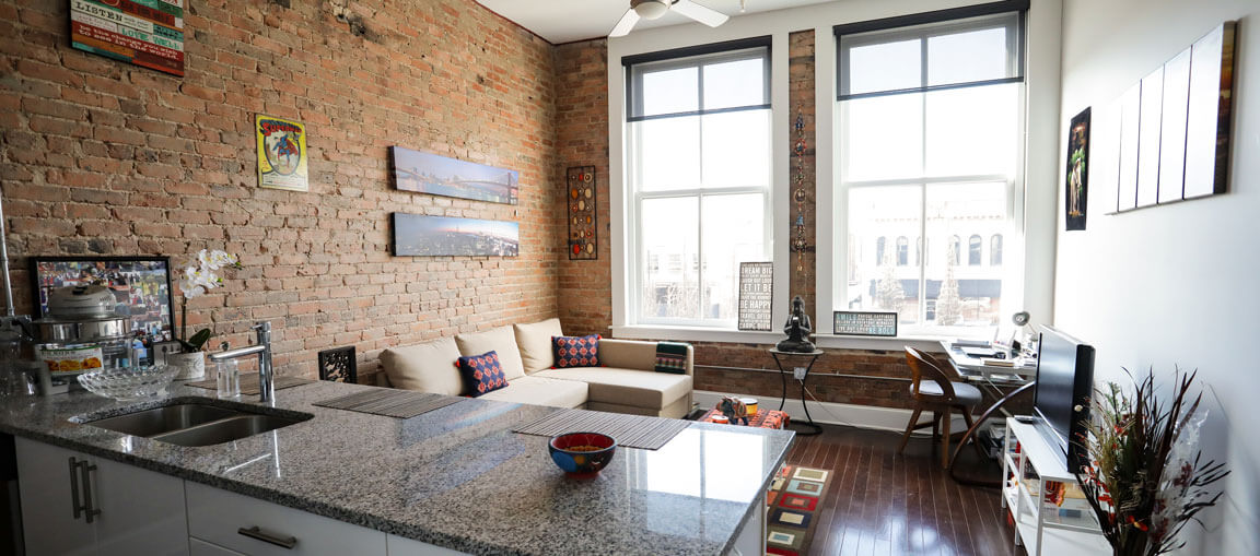 Modern loft with exposed brick walls, big windows, granite kitchen island, beige sectional sofa, desk by window, and wall art.