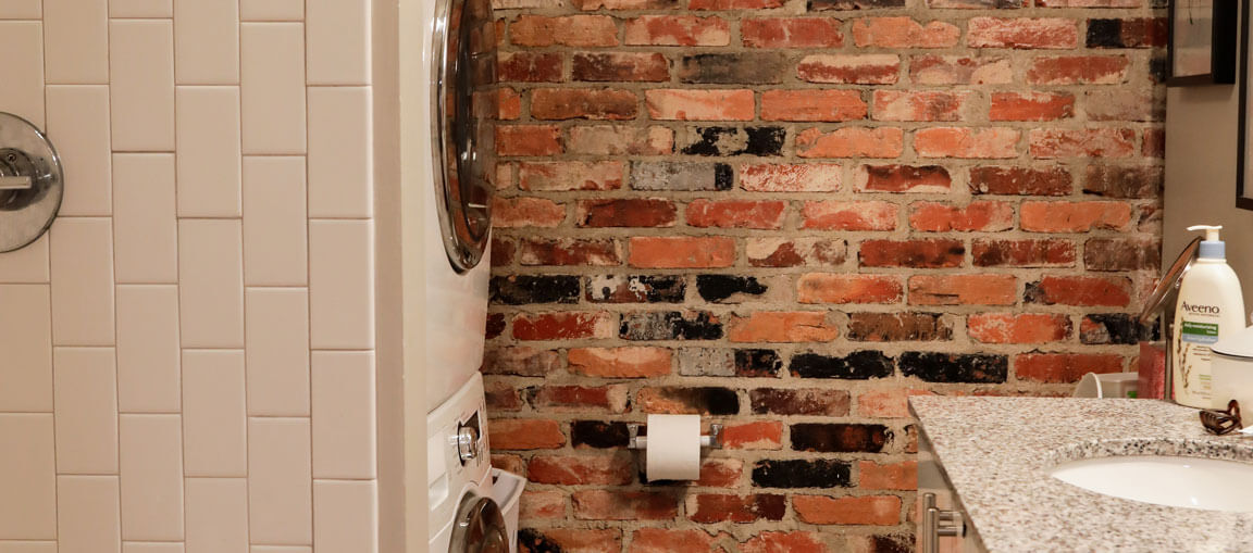 Bathroom featuring a brick accent wall, stacked washer and dryer, wall-mounted toilet paper holder, and granite countertop with soap dispenser.
