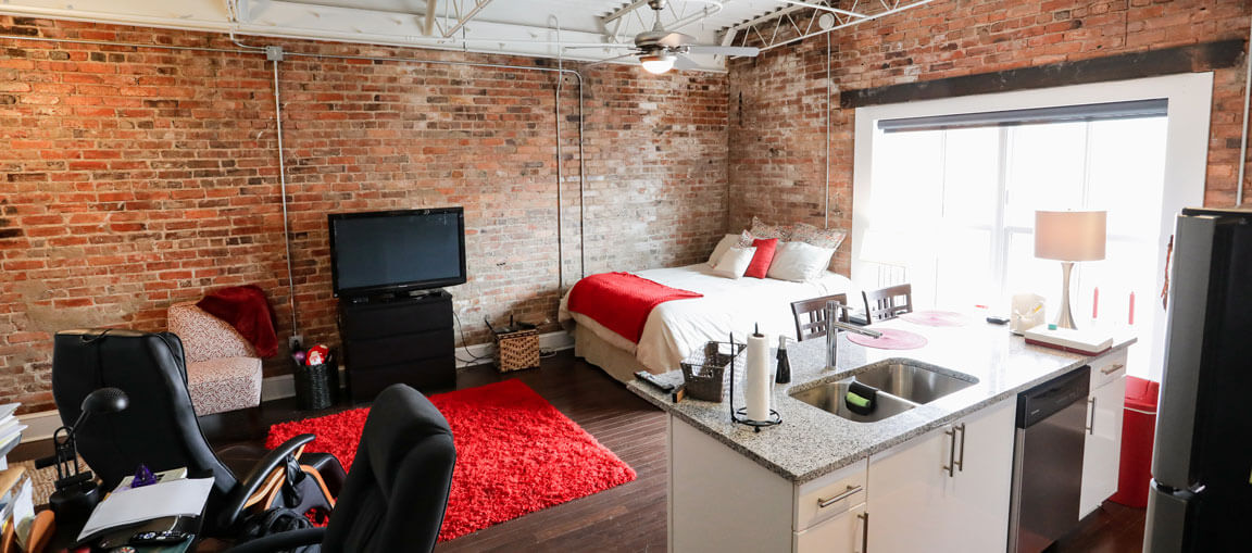 Modern studio apartment with exposed brick, red and white bedding, TV, desk, red rug, white kitchen cabinets, and large windows.