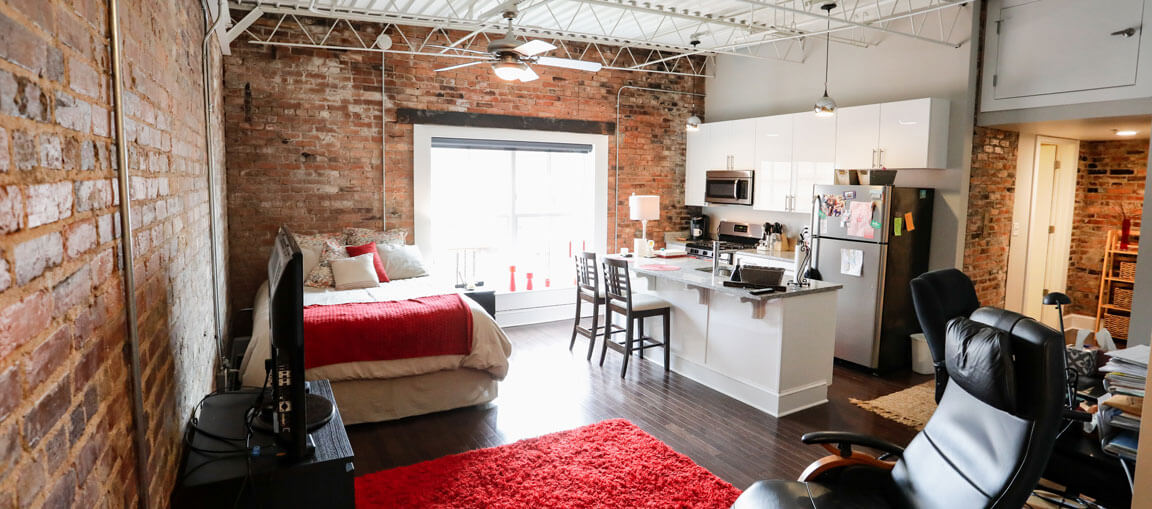 Urban loft with exposed brick, red and white bed, modern kitchen, bar stools, fridge, TV, black office chair, red rug on dark floors.